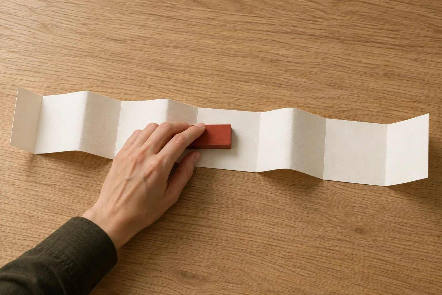 An anonymous hand slides a brick-red block along a blank folded paper strip on an oak table.