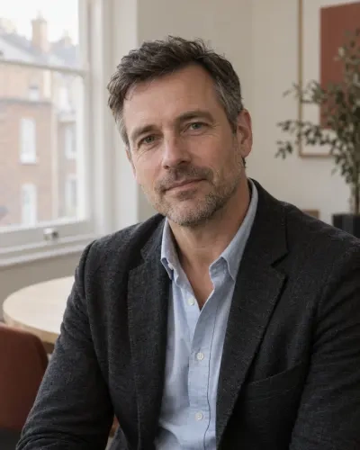Portrait of Rowan Hale: short dark hair greying at the temples, blue shirt and charcoal jacket, seated and slightly turned.